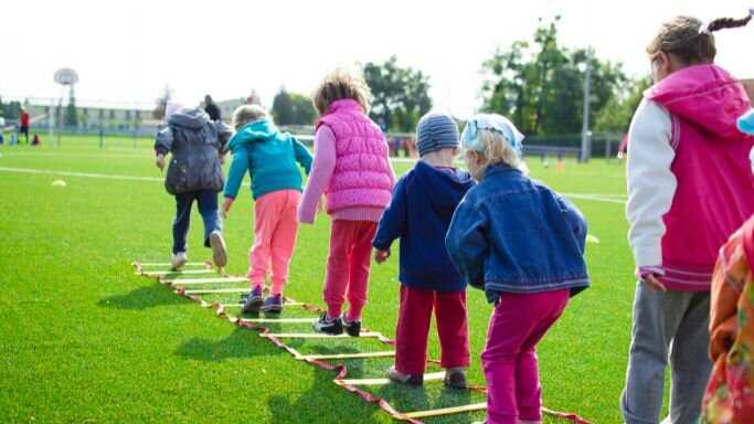 A group of young children jump across a rope ladder on a green field