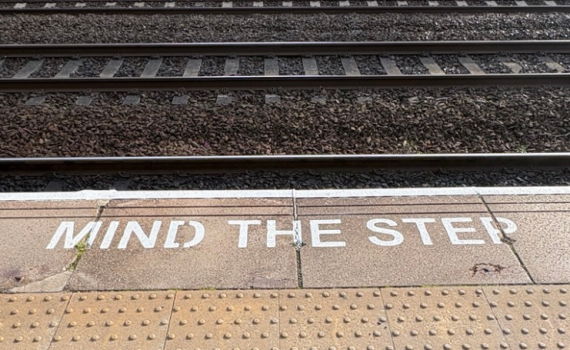 Photograph of a railway platform, 'mind the step' is written at the platform edge in large white capital letters, tracks are visible behind.
