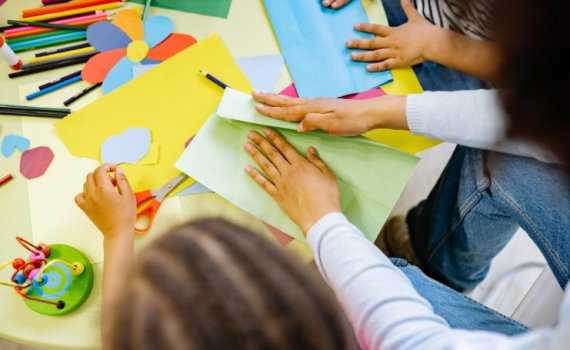 Bird's eye view of a woman's hands folding a piece of pale green card, sat between children at a table with more coloured card, scissors and pencils.