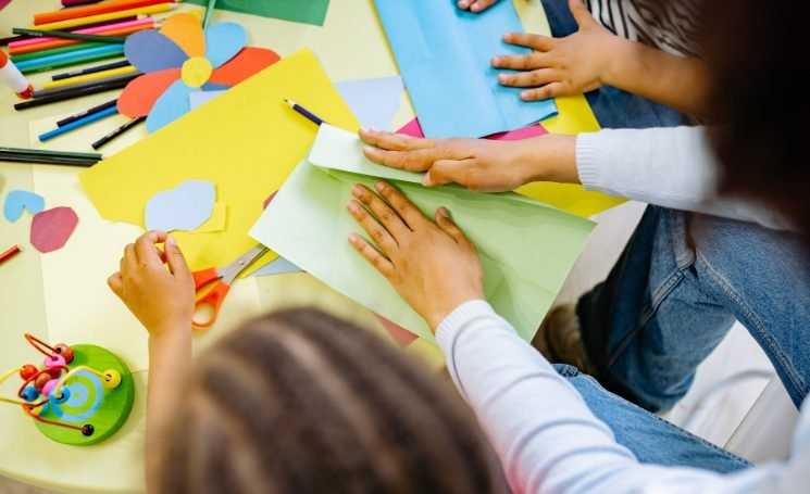 Bird's eye view of a woman's hands folding a piece of pale green card, sat between children at a table with more coloured card, scissors and pencils.